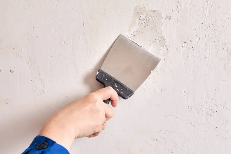 Mano con espátula metálica alisando una pared de yeso con textura irregular.