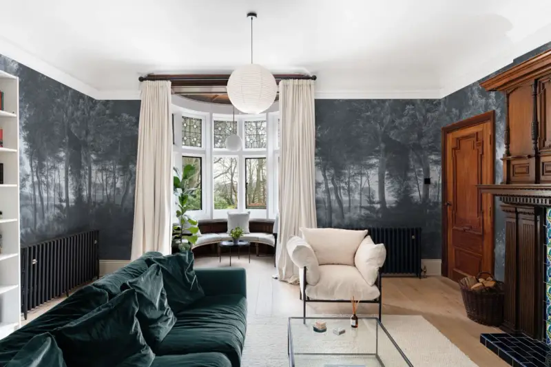 Sala con papel pintado de bosque, sofá verde, sillón beige y chimenea de madera. Ideas de como poner papel pintado para transformar tu hogar.